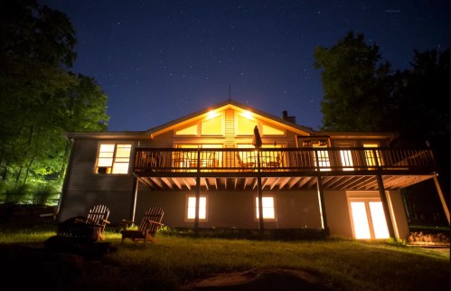 A cozy house at night, illuminated by warm lights, surrounded by trees and a starry sky.