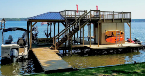 A lakeside dock with a deck, stairs, kayaks, and boats, surrounded by calm water and greenery.