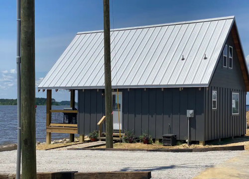 A gray cabin with a metal roof by a lake, featuring a porch and surrounded by gravel and plants.