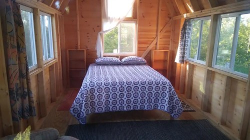 Cozy wooden cabin interior with a bed, patterned blanket, and large windows letting in natural light.
