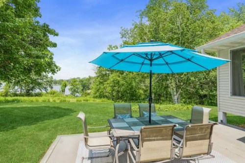 A patio with a glass table and six chairs under a large blue umbrella, surrounded by green grass and trees.