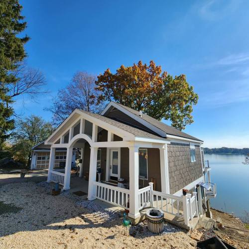 A cozy lakeside house with a porch, surrounded by trees and a clear blue sky, reflecting on the water.
