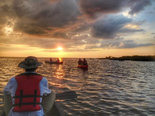 A person in a boat watches as the sun sets over a calm waterway, with other boats in the distance under a cloudy sky.
