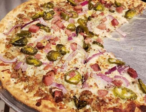 A close-up of a pizza topped with cheese, jalapeños, red onions, and chunks of meat, with a slice removed.