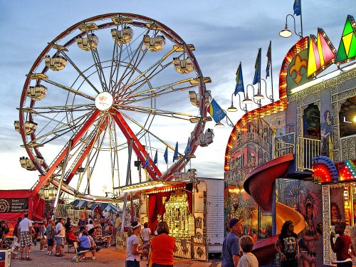 A vibrant carnival scene at dusk featuring a Ferris wheel and colorful game booths with people enjoying the festivities.