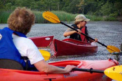 Two people kayaking on a calm lake, surrounded by greenery, with one paddling and the other looking back.