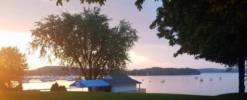 A serene lakeside view at sunset, featuring a tree, a small building, and boats on the water.