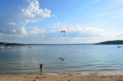 A beach scene with a person standing in shallow water, a paraglider above, and boats on a calm lake under a blue sky.