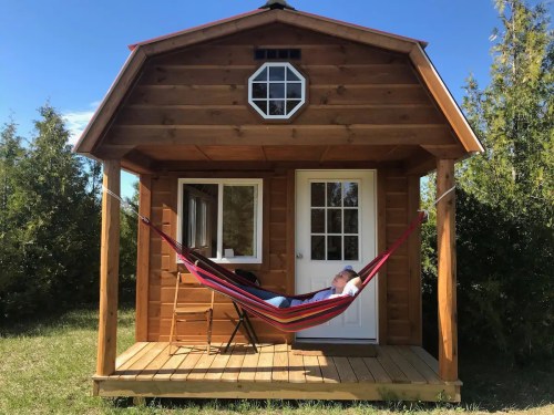 A person relaxes in a hammock on the porch of a small wooden cabin surrounded by greenery.