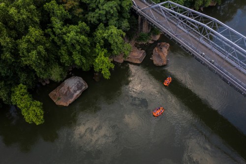 Aerial view of two orange rafts on a river, surrounded by lush greenery and a metal bridge overhead.