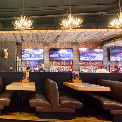 Cozy restaurant interior with wooden decor, antler chandeliers, and multiple TVs showing sports. Booth seating visible.