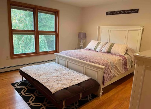 Cozy bedroom featuring a white bed, patterned bedding, a bench, and large windows with natural light.