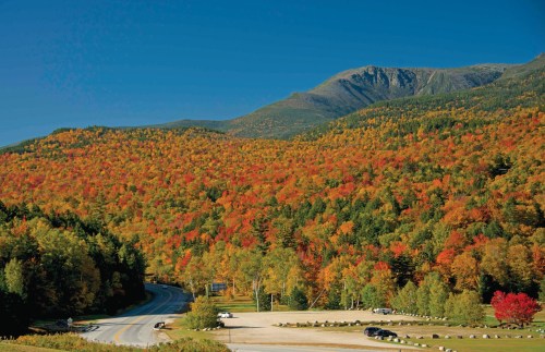 Vibrant autumn foliage covers rolling hills under a clear blue sky, with a winding road and parking area in the foreground.