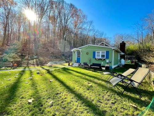 A cozy green house surrounded by trees, with sunlight shining and two lounge chairs on the grassy yard.