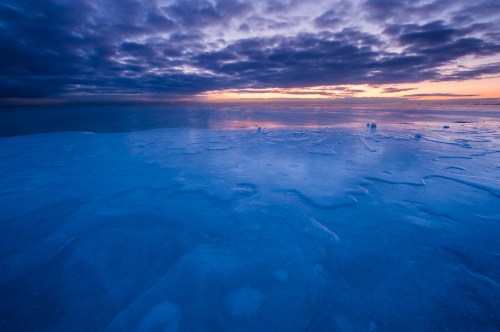 A serene icy landscape at sunset, with a blue-tinted surface reflecting clouds and warm hues in the sky.