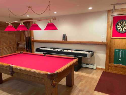 A cozy game room featuring a red pool table, air hockey table, and dartboard with red lighting.