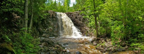 A serene waterfall cascades over rocks, surrounded by lush green trees and foliage in a tranquil forest setting.