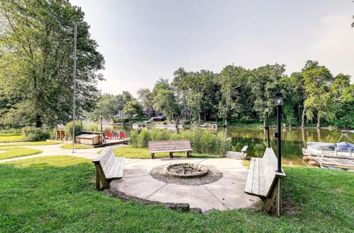 A serene riverside scene featuring a fire pit surrounded by benches, with boats and lush greenery in the background.