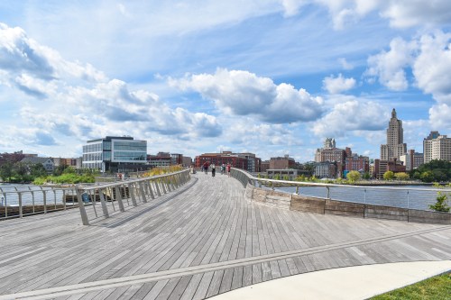 A wooden walkway extends over water, with people walking and a city skyline under a blue sky with fluffy clouds.