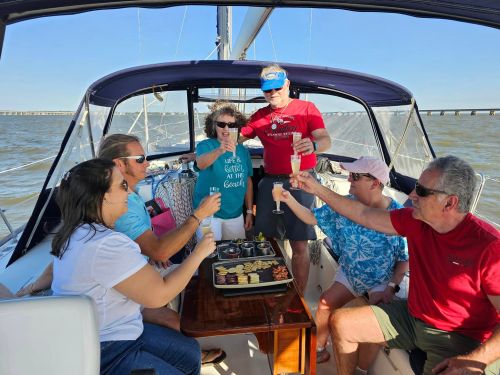 A group of six people on a sailboat, raising drinks and enjoying snacks under a clear blue sky.
