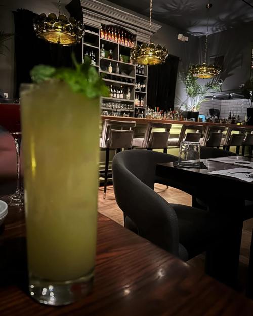 A refreshing cocktail with mint sits on a wooden table, with a stylish bar and seating in the background.