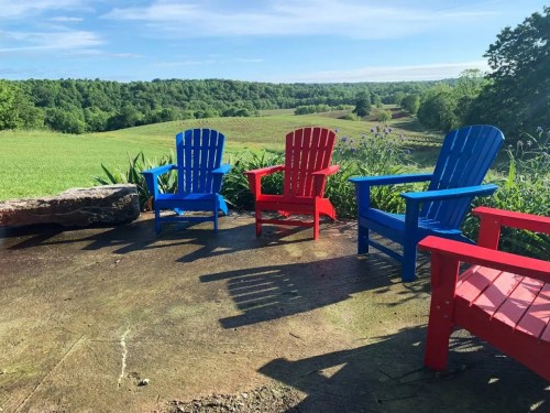 Four colorful Adirondack chairs (two blue, one red) sit on a patio overlooking a lush green landscape under a clear sky.