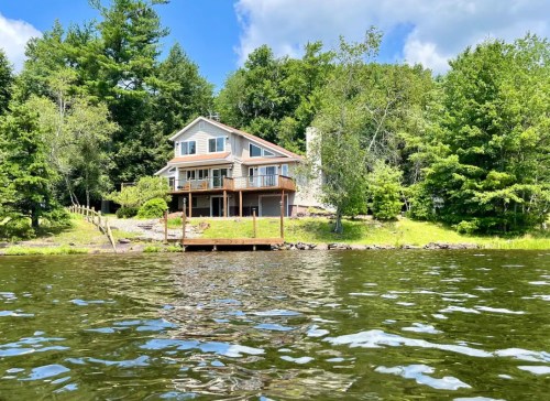 A serene lakeside house surrounded by trees, with a wooden deck and calm water in the foreground.