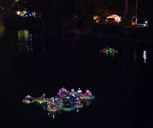 A group of people on illuminated rafts in a dark lake, surrounded by trees and distant camp lights.
