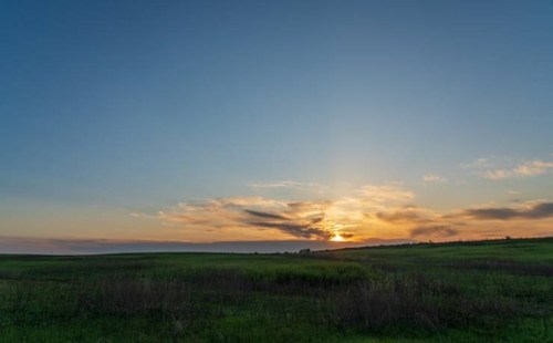 A serene landscape at sunset, featuring a vast green field under a colorful sky with soft clouds.