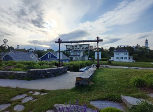 A sign reading "Thank You, Please Come Again" in a scenic area with houses and a cloudy sky in the background.