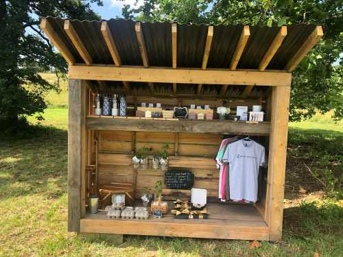 A wooden stall with a corrugated roof, displaying various products like shirts, jars, and plants on shelves.