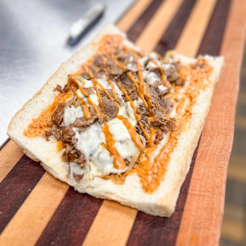 A sandwich on a wooden board, filled with sliced beef, melted cheese, and drizzled with sauce.