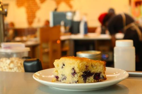 A slice of blueberry cake on a white plate, with a blurred café background.