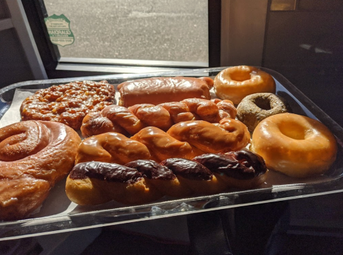 A tray of assorted donuts, including glazed, chocolate-covered, and cinnamon rolls, displayed in natural light.