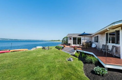 A scenic waterfront view of a house with a deck, lush green lawn, and kayaks along the shore under a clear blue sky.