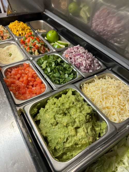 A variety of fresh toppings in metal trays, including guacamole, diced tomatoes, onions, cilantro, cheese, and lime.