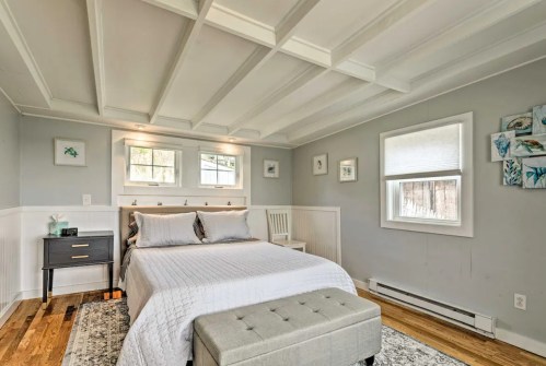 Cozy bedroom with a white bed, gray accents, wooden floors, and natural light from windows.