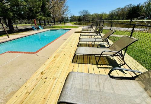 A sunny pool area with lounge chairs and a clear blue pool, surrounded by green trees and a fence.
