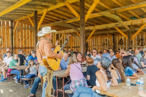 A musician performs with a guitar in a rustic barn, entertaining a large audience seated at wooden tables.