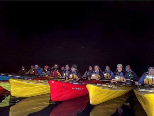 A group of kayakers in brightly colored kayaks paddling at night, illuminated by soft light on the water.