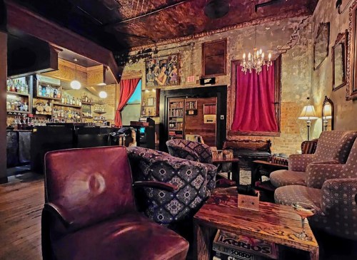 Cozy bar interior with vintage decor, plush seating, a bookshelf, and a warm ambiance created by soft lighting.