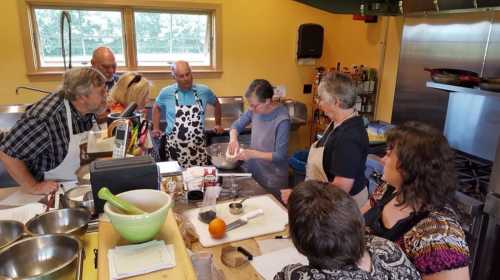 A group of people in an inviting kitchen, gathered around a table, watching a woman mix ingredients in a bowl.