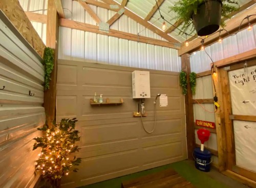 A rustic outdoor shower area with a water heater, plants, and decorative lights, featuring a urinal and wooden bench.