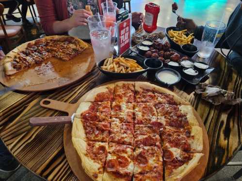 A table with two pizzas, one with pepperoni, and a platter of fries and chicken wings with dipping sauces.