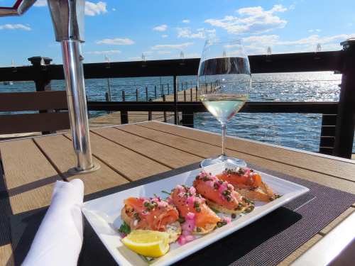 A plate of salmon appetizers with lemon, paired with a glass of white wine, overlooking a serene waterfront.