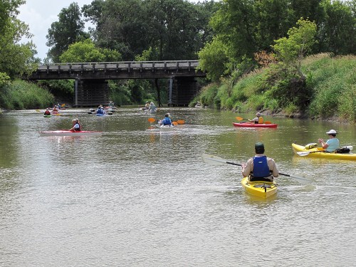 A group of kayakers paddles down a calm river under a bridge, surrounded by lush greenery.