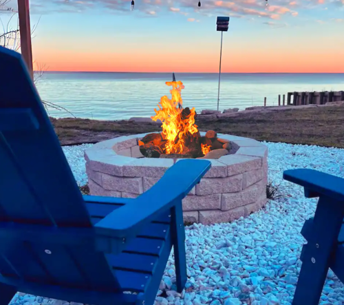 A cozy fire pit surrounded by blue chairs, with a serene lake and sunset in the background.