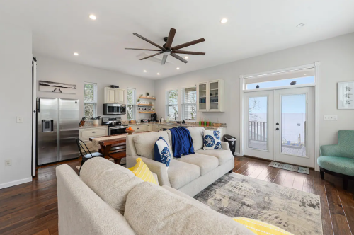 Bright, modern living space with a kitchen, cozy sofa, and large windows overlooking a deck and water view.