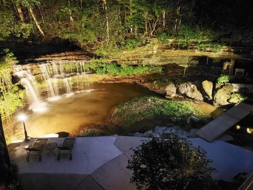 A serene nighttime scene of a waterfall cascading into a pond, surrounded by lush greenery and a stone pathway.
