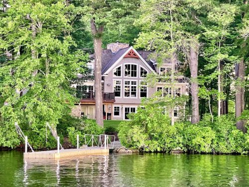 A large, modern house surrounded by trees, overlooking a calm lake with a small dock in the foreground.